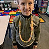 boy, candy_necklace, child, classroom, clothing, colorful, concrete_floor, educational_rug, eyes, face, hair, indoor, learning_environment, person, shelves, smiling, standing, table, young, zipper_jacket