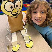 Koltyn joined the competition — help win amazing prizes! arts_and_crafts, brown_paper_bag, cartoon_face, child, classroom, colorful, creative, curly_hair, cut_out, educational_materials, happy, heart_shape, indoor, kid, paper_craft, paper_legs, person, project, smiling, table