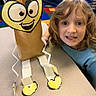 arts_and_crafts, brown_paper_bag, cartoon_face, child, classroom, colorful, creative, curly_hair, cut_out, educational_materials, happy, heart_shape, indoor, kid, paper_craft, paper_legs, person, project, smiling, table