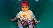 Michael is registered to the contest to win money with this photo: casual_clothing, child, daylight, fun, green_slide, happy, outdoor, person, playground, recreation, red_cap, shorts, sitting, slide, smiling, socks, toddler, trees, white_tshirt, wood_chips