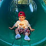 casual_clothing, child, daylight, fun, green_slide, happy, outdoor, person, playground, recreation, red_cap, shorts, sitting, slide, smiling, socks, toddler, trees, white_tshirt, wood_chips