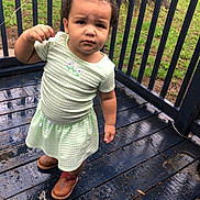 Braelynn is registered to the contest to win money with this photo: toddler, child, outdoor, porch, wooden_floor, rain, wet_surface, curly_hair, topknot, green_dress, brown_boots, expression, serious_face, grass, railing, tree_trunk, nature, daylight, person, standing
