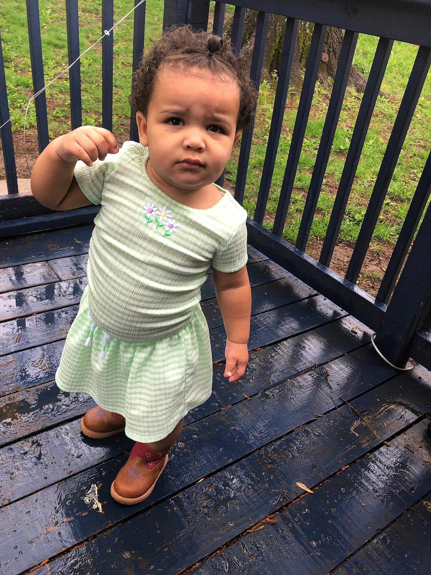 Braelynn is registered to the contest to win money with this photo: toddler, child, outdoor, porch, wooden_floor, rain, wet_surface, curly_hair, topknot, green_dress, brown_boots, expression, serious_face, grass, railing, tree_trunk, nature, daylight, person, standing