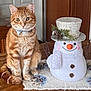 cat, orange_tabby, bow_tie, pet, indoor, table, wooden_table, snowman, winter_decoration, hat, pinecones, twigs, white_fur, orange_nose, smile, decorative, holiday, cozy, curious, domestic_animal