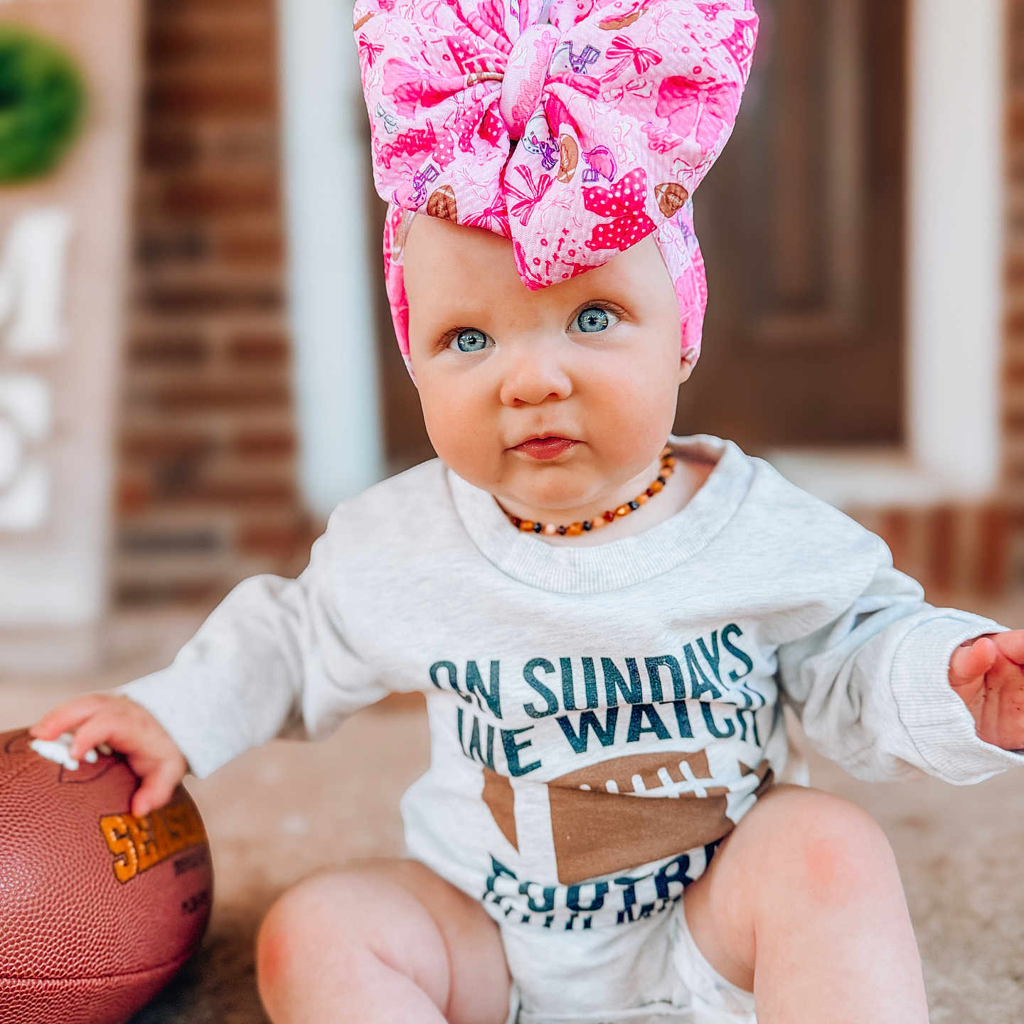 Sutton joined the competition — help win amazing prizes! baby, child, infant, pink_bow, headwrap, blue_eyes, onesie, football, porch, brick, sitting, cute, toddler, outdoor, portrait, clothing, hand, face, expression, casual