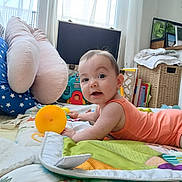 Héléna a rejoint le concours — aidez-le/la à gagner de superbes lots ! baby, child, infant, play_mat, toy, orange_onesie, cushion, indoor, curious, face, expression, furniture, books, blanket, window, daylight, floor, room, smiling, cute