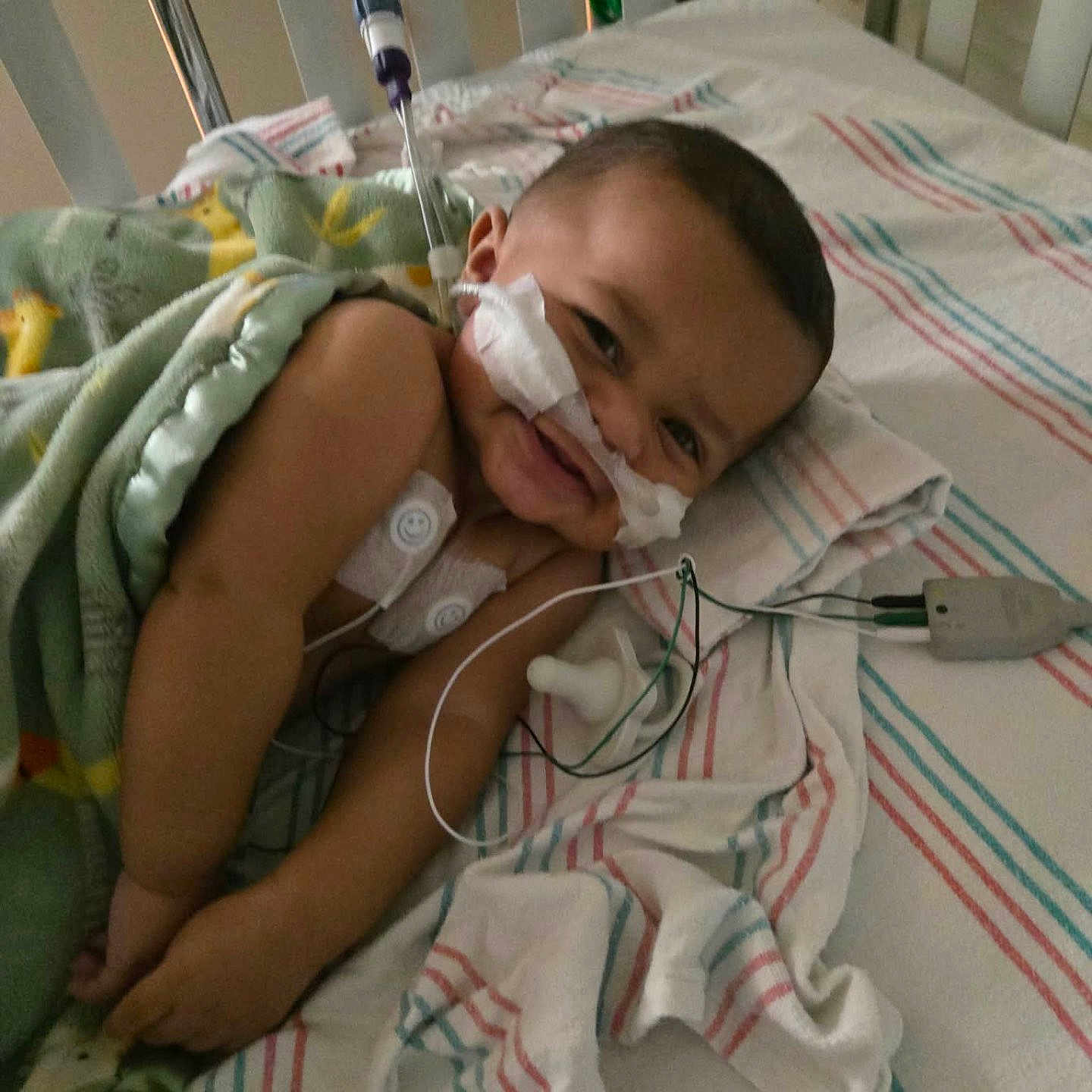 Veylin is registered to the contest to win money with this photo: baby, hospital, crib, medical_tubes, blanket, smile, face, child, monitoring_wires, striped_sheet, healthcare, lying_down, cute, infant, toddler, medical_equipment, cozy, child_patient, happy, hospital_bed