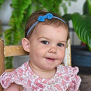 Kylie joined the competition — help win amazing prizes! toddler, child, smiling, headband, blue_bow, pink_dress, patterned_clothing, wooden_chair, green_plants, indoor, portrait, cute, happy, face, person, baby, short_hair, candid, seated, natural_light