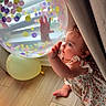 baby, child, balloon, confetti, floor, curtain, dress, floral_pattern, hand, foot, indoor, natural_light, colorful, transparent, cute, curious, sitting, infant, playful, soft_light
