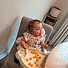 toddler, child, high_chair, snack, cheese_puffs, floral_dress, purple_bow, eating, indoor, table, chair, carpet, curtains, wall, foot, cute, baby_food, person, dining_room, happy