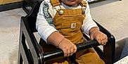 Kyzeir Walker is registered to the contest to win money with this photo: toddler, child, high_chair, overalls, curly_hair, indoor, kitchen, table, curious, sitting, safety_bar, floor, wood_paneling, boxes, shelf, white_shirt, person, young_child, face, expression