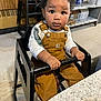 toddler, child, high_chair, overalls, curly_hair, indoor, kitchen, table, curious, sitting, safety_bar, floor, wood_paneling, boxes, shelf, white_shirt, person, young_child, face, expression