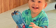 Mario joined the competition — help win amazing prizes! toddler, child, smiling, curly_hair, plastic_bottle, green_shirt, shark_pattern, gray_shorts, sitting, floor, indoor, happy, cute, playful, person, young_child, bright, casual_clothing, baby, portrait