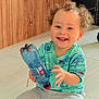 toddler, child, smiling, curly_hair, plastic_bottle, green_shirt, shark_pattern, gray_shorts, sitting, floor, indoor, happy, cute, playful, person, young_child, bright, casual_clothing, baby, portrait