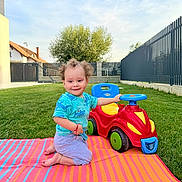 Mario joined the competition — help win amazing prizes! toddler, child, curly_hair, toy_car, outdoor, grass, blue_sky, striped_mat, play, smile, kneeling, house, fence, greenery, daylight, casual_clothing, happy, backyard, person, summer