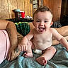 Letty is registered to the contest to win money with this photo: baby, smiling, tongue_out, curly_hair, diaper, blanket, tattoo, adult, indoor, bed, wooden_wall, door, plaid_shorts, person, happy, skin, foot, hand, furniture, room