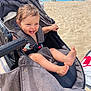 toddler, stroller, beach, sand, smiling, sunny, child, feet, pacifier, gray_fabric, outdoor, happy, playful, baby, vacation, summer, person, seat, relaxing, sunlight