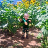 baby, blue_sky, boots, child, dirt, farmland, flowers, football_clothing, garden, green_shirt, greenery, happy, nature, outdoor, patterned_pants, portrait, standing, sunflower_field, sunflowers, sunlight