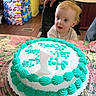 baby, cake, birthday_cake, candle, high_chair, kitchen, child, icing, number_candle, table, curious, celebration, person, food, indoor, hand, frosting, plastic_container, decorated_cake, young_child