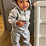 toddler, child, hoodie, sweatpants, sneakers, standing, smiling, indoor, wooden_floor, storage_unit, grey_clothing, pink_detail, casual_wear, happy, person, young_child, cute, playful, holding_object, expression