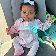 Tutu is registered to the contest to win money with this photo: baby, child, car_seat, onesie, pink_clothing, white_bow, curly_hair, infant, seatbelt, car_interior, cute, portrait, sitting, young_child, face, looking, sunlight, clothing, hand, footed_pajamas