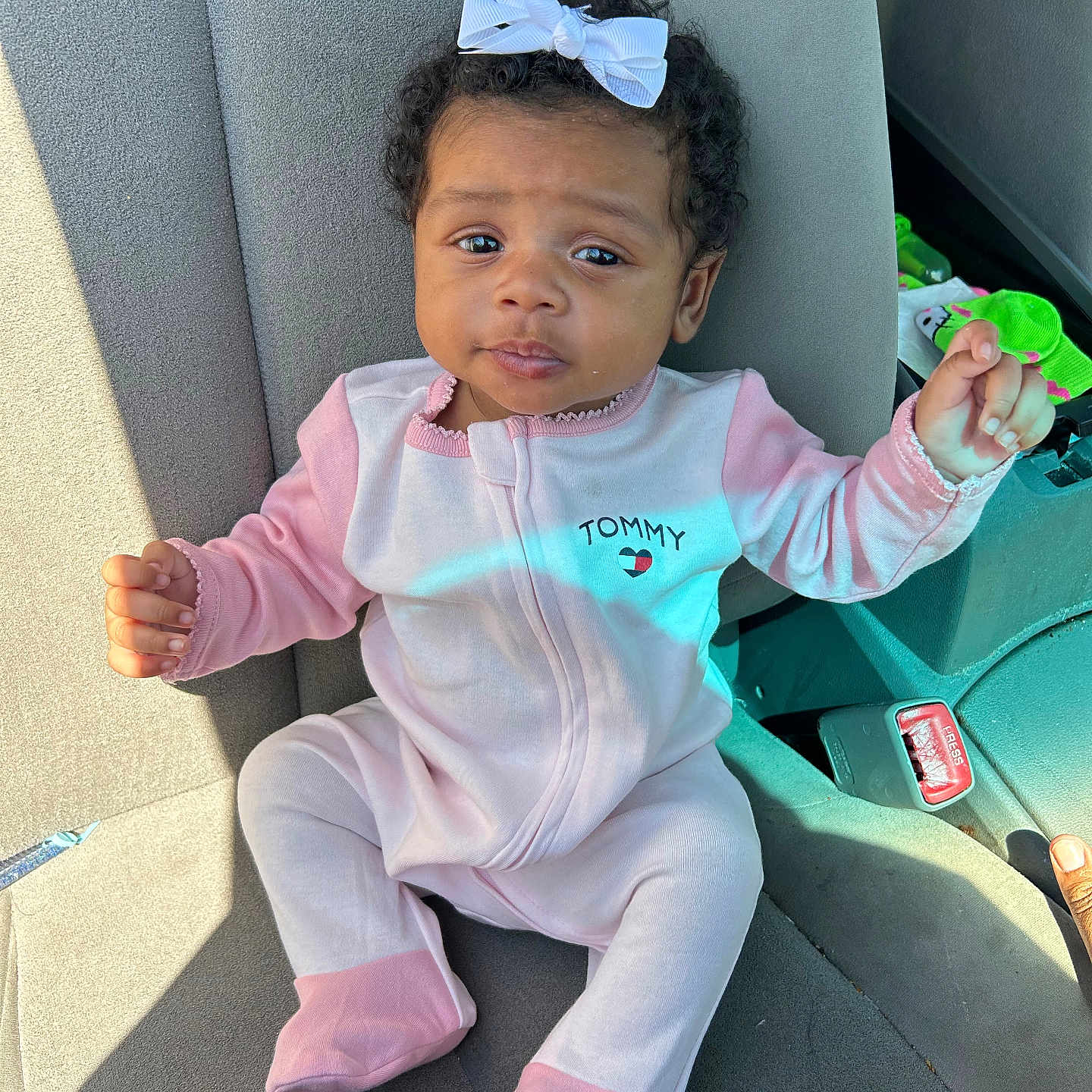 Tutu is registered to the contest to win money with this photo: baby, child, car_seat, onesie, pink_clothing, white_bow, curly_hair, infant, seatbelt, car_interior, cute, portrait, sitting, young_child, face, looking, sunlight, clothing, hand, footed_pajamas