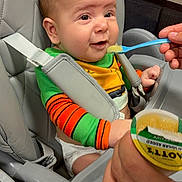 Kaige is registered to the contest to win money with this photo: adult_hand, baby, bib, child, eating, feeding, food, green_clothing, happy, high_chair, indoor, infant, kitchen, mealtime, orange_clothing, portrait, safety_harness, seated, smiling, spoon