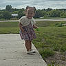 toddler, child, smiling, outdoor, sidewalk, grass, toy, dress, white_shoes, pigtails, cloudy_sky, greenery, young_child, person, nature, playful, happy, casual_clothing, daytime, small_toy