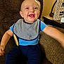baby, child, smiling, blue_clothing, striped_bib, sitting, arm_outstretched, brown_chair, indoor, happy, face, person, cute, young_child, home_interior, wooden_table, decor, teeth, portrait, infant