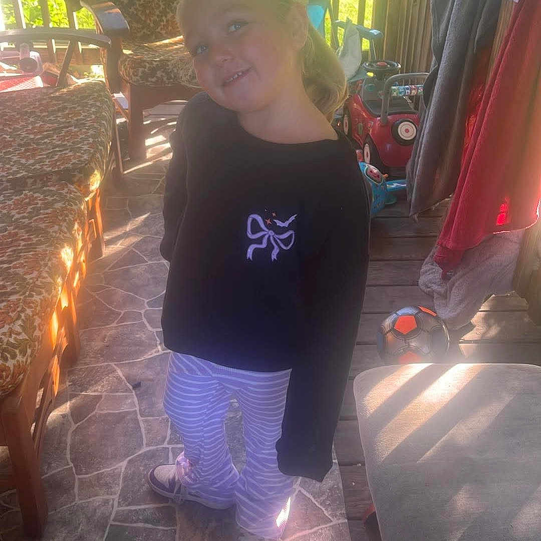 Phoebe is registered to the contest to win money with this photo: child, girl, smiling, portrait, standing, outdoor, porch, sunlight, patio, chair, clothing, toy, soccer_ball, deck, railing, garden, house, striped_pants, black_shirt, happy