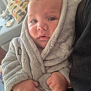 Dylan is registered to the contest to win money with this photo: baby, child, closeup, clothing, cozy, cushions, cute, eyes, face, fists, fluffy, hands, indoors, infant, living_room, pattern, person, portrait, robe, soft