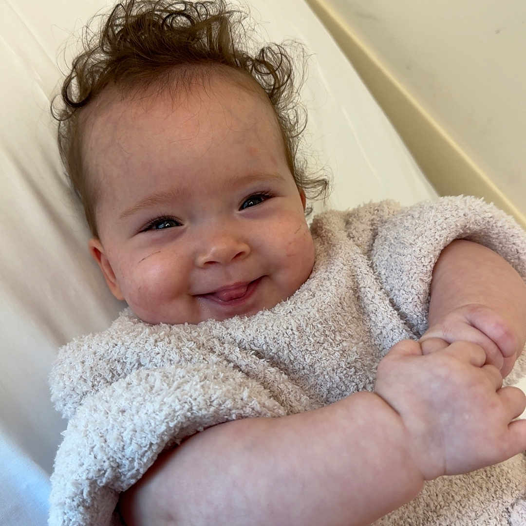 Alice Ophelia is registered to the contest to win money with this photo: baby, child, smiling, curly_hair, towel, blanket, skin, face, happy, infant, cozy, cute, lying_down, indoors, person, portrait, young_child, cheeks, hands, relaxed