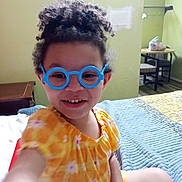 Kai'Lani Garnett is registered to the contest to win money with this photo: child, smiling, curly_hair, glasses, toy_glasses, yellow_dress, floral_pattern, bed, quilt, indoor, room, table, chair, furniture, person, happy, portrait, casual, playful, young