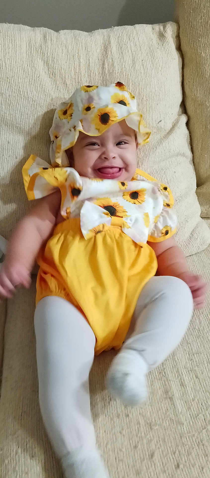 Astrid is registered to the contest to win money with this photo: baby, child, smiling, sunflower, hat, yellow_clothing, white_tights, couch, indoors, happy, person, cute, infant, portrait, sitting, furniture, cozy, playful, tongue_out, fabric