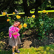 Eva is registered to the contest to win money with this photo: child, girl, yellow_roses, flower_bush, plush_toy, pink_toy, leopard_print_dress, sandals, park, tree, grass, sunlight, shadow, nature, outdoor, smiling, happy, greenery, bench, people