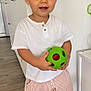 Marceau participe au concours pour gagner de l'argent avec cette photo : toddler, child, toy, green_ball, barefoot, striped_pants, white_shirt, indoor, flooring, kitchen, modern, curly_hair, cute, smiling, standing, home, play, person, young_child, casual_clothing