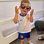 toddler, child, sunglasses, sport_outfit, indoor, crib, floor, tile_floor, shorts, shirt, socks, person, playful, fashion, pose, cute, home, smile, hair, footwear