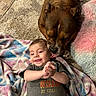 baby, baby_clothing, blanket, brown_dog, carpet, cuddling, dog, family, friendship, happy, home, indoor, infant, lying_down, pet, pink_pillow, portrait, short_hair, smiling, text_on_shirt