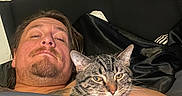 Mongol And Shotgun is registered to the contest to win money with this photo: man, cat, bed, blanket, tabby_cat, person, indoor, resting, relaxed, sleepy, headboard, furniture, animal, pet, comfort, portrait, closeup, face, human, cozy