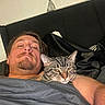 man, cat, bed, blanket, tabby_cat, person, indoor, resting, relaxed, sleepy, headboard, furniture, animal, pet, comfort, portrait, closeup, face, human, cozy