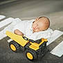 baby, sleeping, blanket, toy_truck, yellow, road, sidewalk, crosswalk, child, infant, cute, peaceful, vehicle, outdoor, small, resting, soft, head, face, wheels