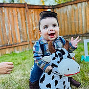 Noah is registered to the contest to win money with this photo: toddler, child, cowboy, plush_toy, cow, boots, hat, outdoor, grass, fence, smiling, happy, playing, person, toy, clothing, baby, cute, fun, daytime
