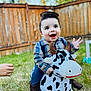 toddler, child, cowboy, plush_toy, cow, boots, hat, outdoor, grass, fence, smiling, happy, playing, person, toy, clothing, baby, cute, fun, daytime