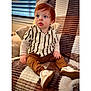 child, toddler, redhead, striped_shirt, sneakers, sitting, blanket, pillow, indoor, window_blinds, portrait, curious_expression, hands, legs, cozy, fashionable, soft_texture, home, casual_clothing, young