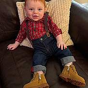 Santino is registered to the contest to win money with this photo: baby, red_hair, plaid_shirt, suspenders, jeans, boots, couch, pillow, smiling, child, indoor, comfortable, casual_clothing, rolled_jeans, footwear, happy, portrait, seated, cute, cozy