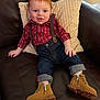 baby, red_hair, plaid_shirt, suspenders, jeans, boots, couch, pillow, smiling, child, indoor, comfortable, casual_clothing, rolled_jeans, footwear, happy, portrait, seated, cute, cozy