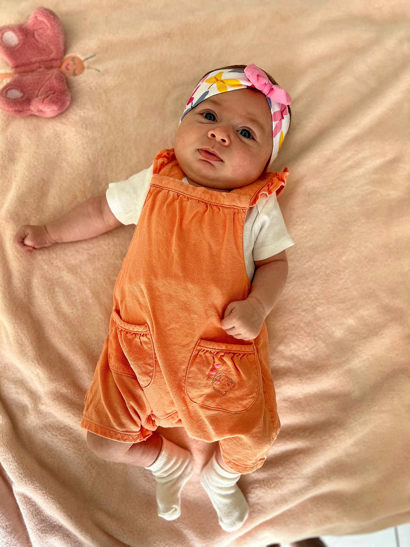 Leyla a rejoint le concours — aidez-le/la à gagner de superbes lots ! baby, infant, headband, orange_overalls, white_shirt, white_socks, blanket, plush_toy, butterfly_toy, soft_texture, lying_down, cute, portrait, child, indoors, cozy, comfortable, small_hands, curious_look, pink