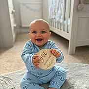 Rowen is registered to the contest to win money with this photo: baby, infant, smiling, blue_onesie, wooden_plaque, eight_months, rug, nursery, crib, carpet, happy, child, indoor, cute, sitting, person, home, soft_lighting, portrait, teeth