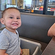 Noah is registered to the contest to win money with this photo: toddler, child, high_chair, smiling, food, indoor, restaurant, table, seat, casual, menu, reflection, window, shorts, shirt, person, happy, baby, cute, dining