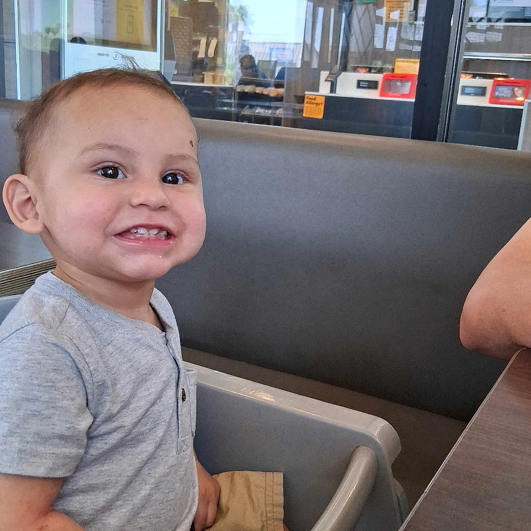 Noah is registered to the contest to win money with this photo: baby, casual, child, cute, dining, food, happy, high_chair, indoor, menu, person, reflection, restaurant, seat, shirt, shorts, smiling, table, toddler, window